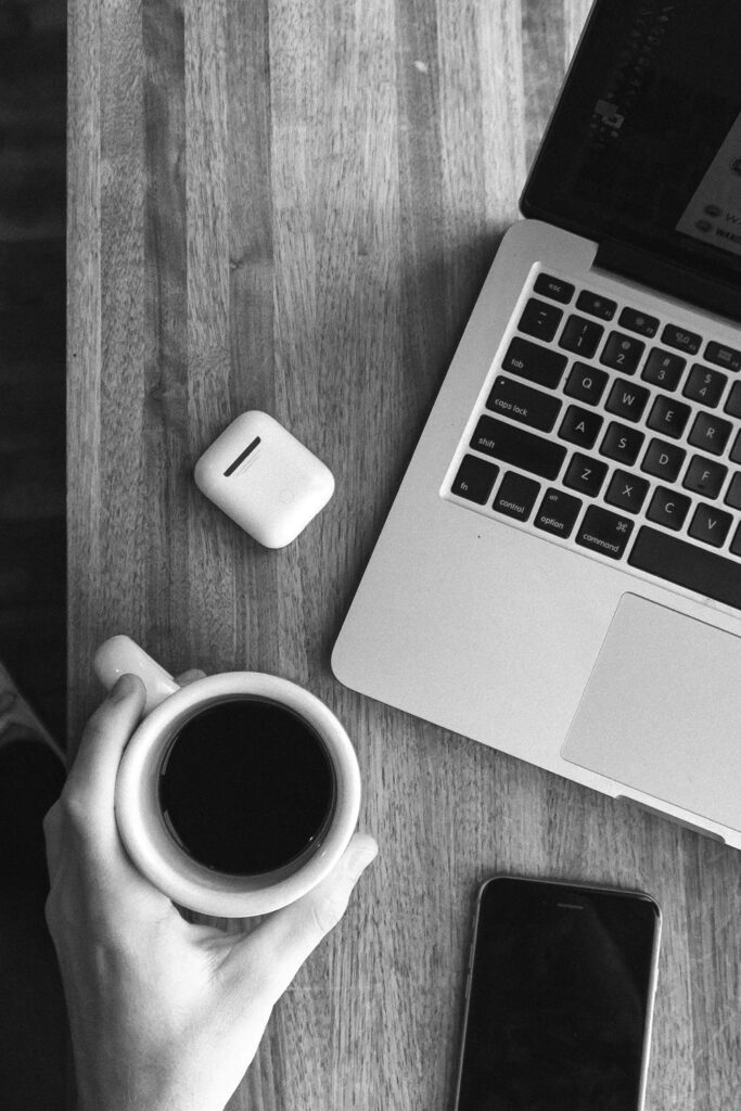 Hand on a desk holding a cup of coffee, surrounded by headphones, a laptop, and a cell phone